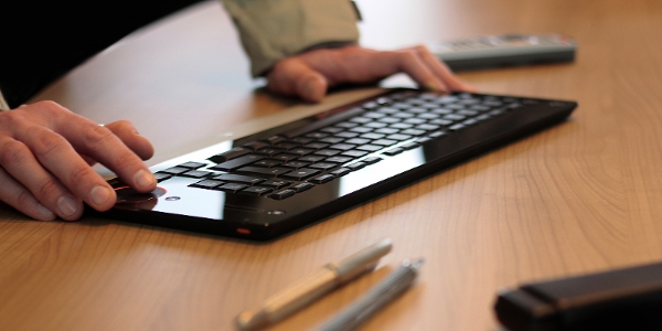 Tippende Hände bedienen eine moderne Funktastatur auf dem Schreibtisch. Stifte und weitere Geräte liegen dabei.