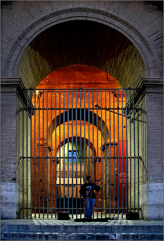 Ein Straßenmusiker mit seiner E-Gitarre vor einem der Bögen des Kolosseums. Die Lichtstimmung ist kühl, innerhalb der Bögen scheint warmes Licht. 