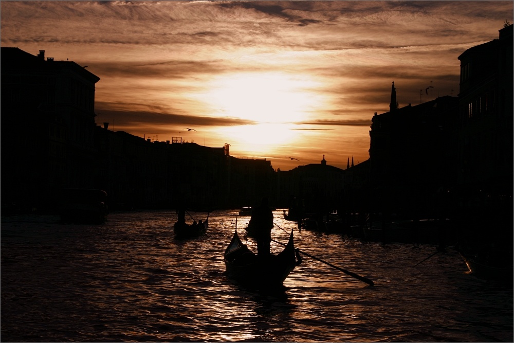 Der Canal Grande bei Sonnenuntergang. Die Sonne spiegelt sich auf dem Wasser. Alle Gebäude und Gondeln sind nur als schwarze Silhouetten zu sehen. 