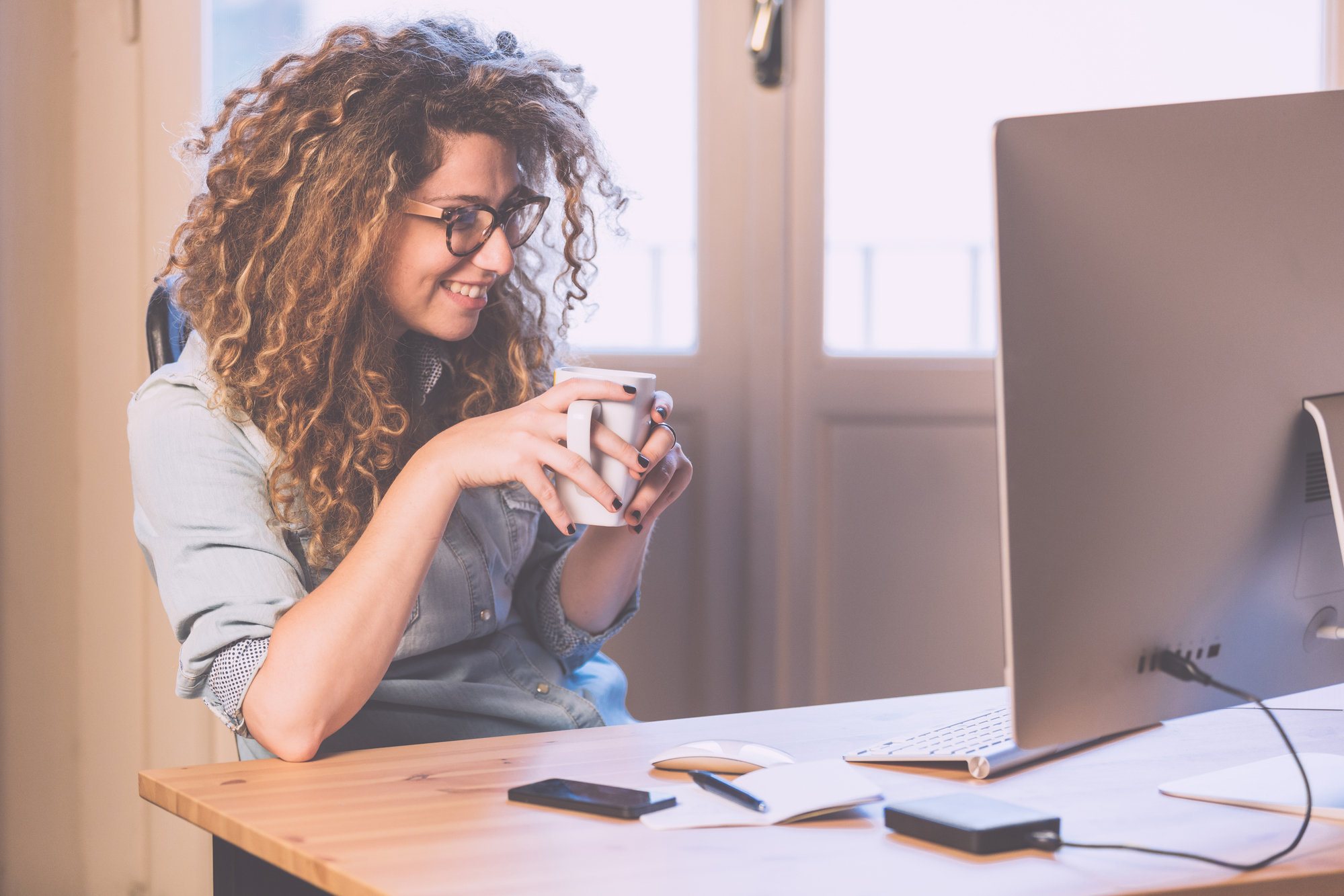 Eine junge Frau sitzt lächelnd vor dem Rechner mit einer Tasse Kaffee in der Hand
